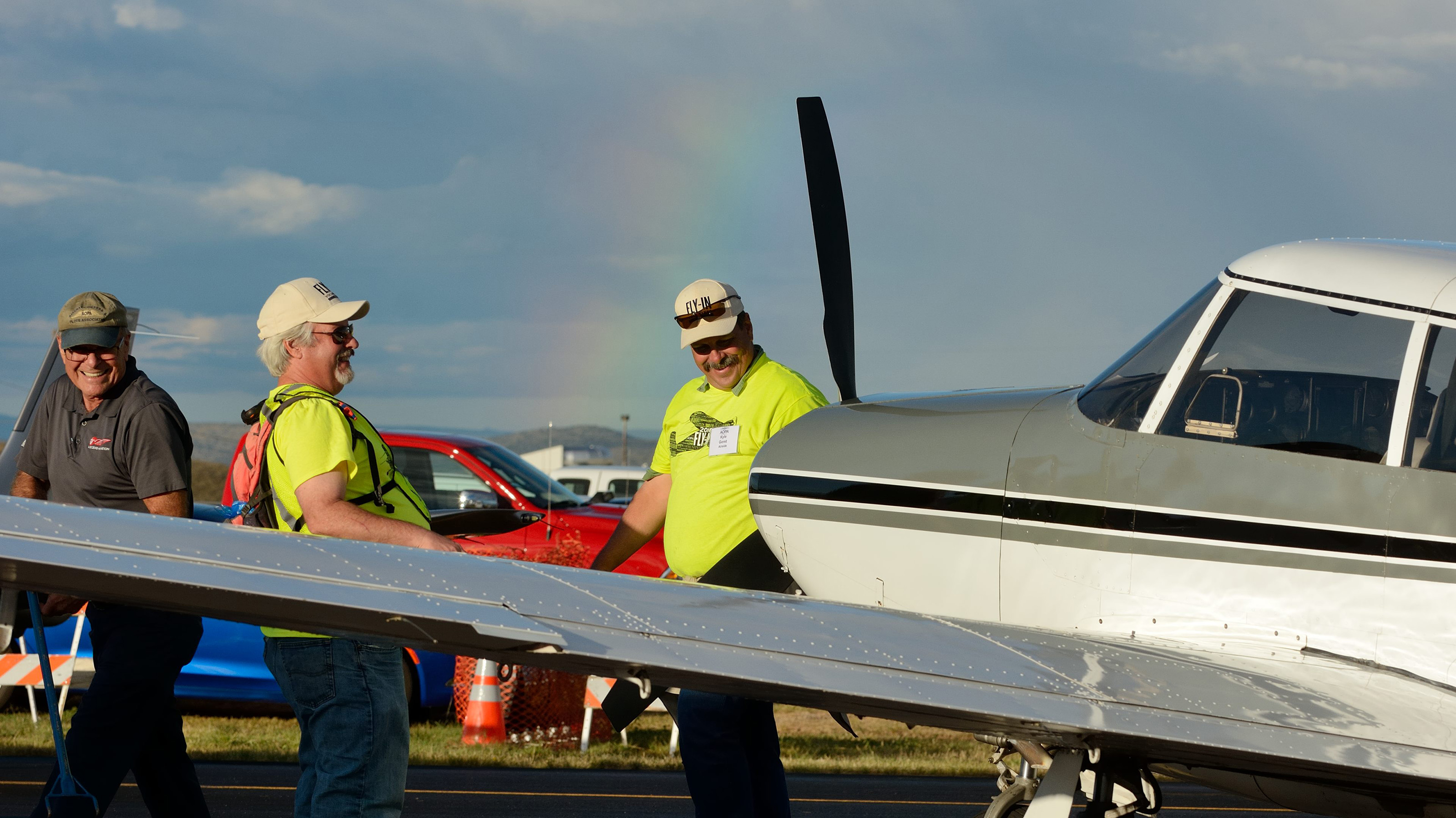 Airside volunteers share a laugh after positioning a Piper Comanche in the static display. A rainbow gives evidence of an afternoon rainshower. Photo by Mike Collins.