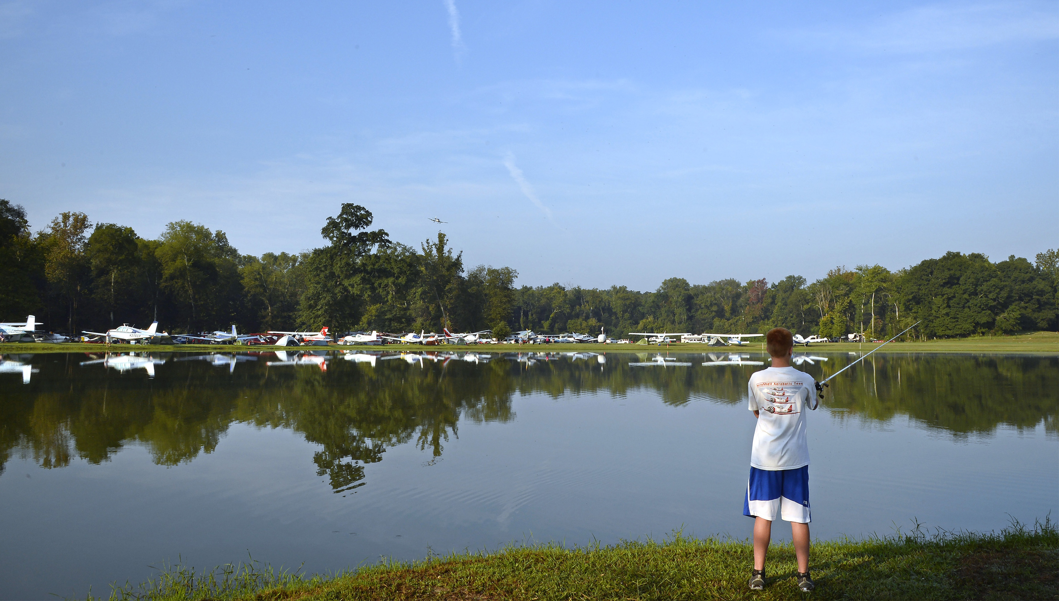 Aircraft are reflected in a pond at Triple Tree Aerodrome where Kyle Carden casts for bass during the annual fly-in. Carden and his father Keith flew from their Irvington, Alabama, home in their Stinson Voyager, an airplane that is now serving the family's fourth generation. Photo by David Tulis.