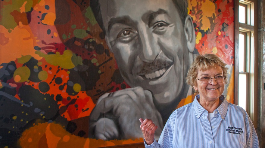 Walt Disney Hometown Museum director Kaye Malins in front of a mural of Walt Disney inside the museum in Marceline, Missouri. Walt says he found his magic as a child growing up in Marceline. Photo by MeLinda Schnyder.