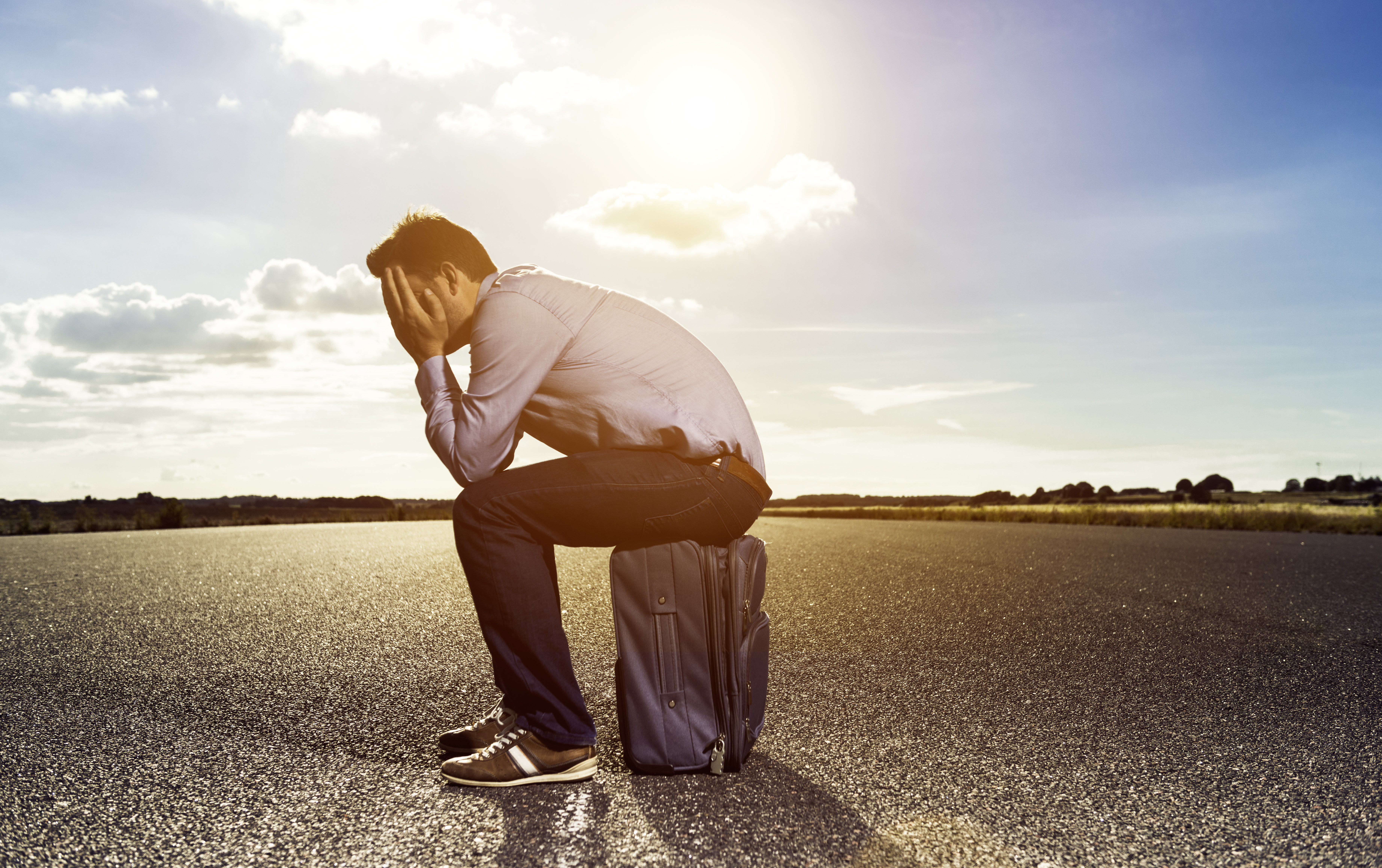 Male passenger sits on a suitcase with his face buried in his hands. He is all alone and waits for a plane to take him home or to take him on a business trip or vacation. He looks bored as he is stranded on the airfield with no hope for take off soon.