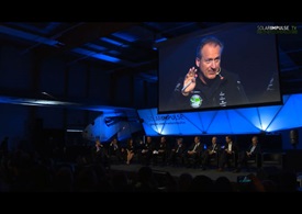 Solar Impulse CEO and pilot André Borschberg (on screen) addresses the crowd gathered for the unveiling of Solar Impulse 2, flanked on stage by project sponsors with the aircraft in the background. 